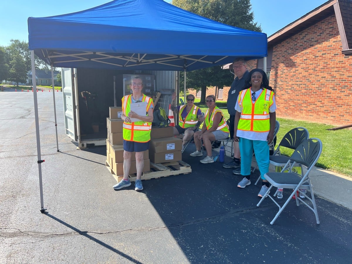 Volunteers in safety vests under a canopy beside a storage container stacked with boxed meals at a 2026 summer pickup site